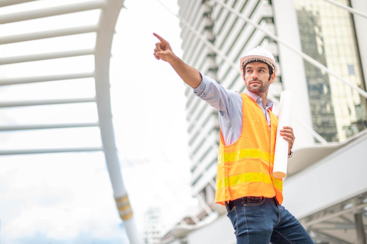 Young caucasian man holding a big paper, guy wearing light blue shirt and jeans with orange vest and white helmet for security in construction area. photo