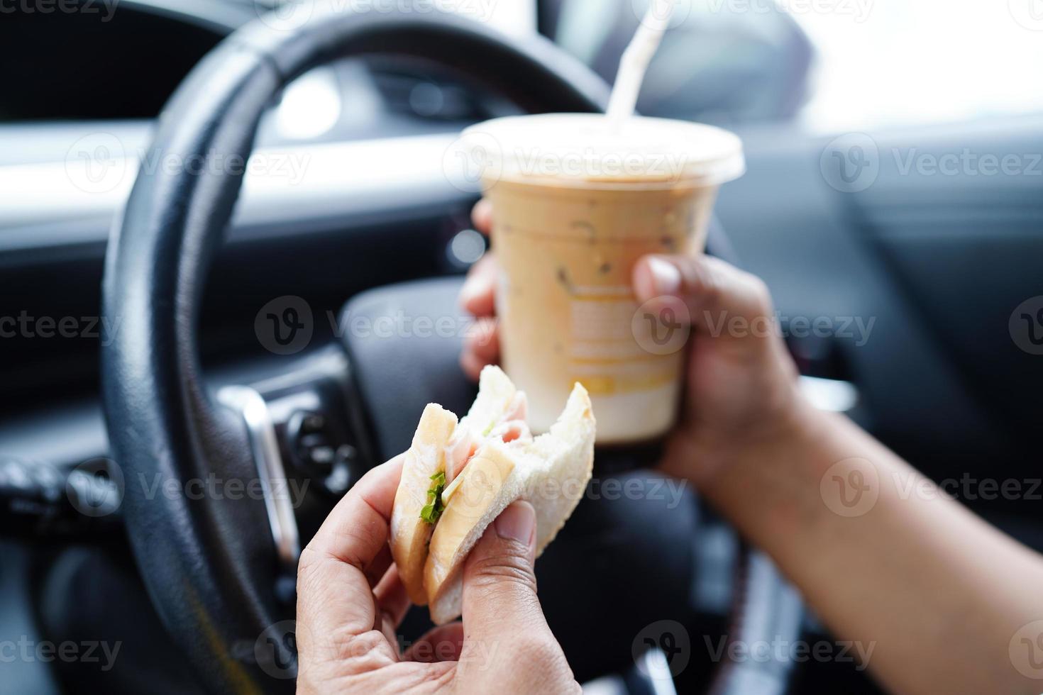 Asian woman driver hold ice coffee cup and sandwich bread for eat and drink in car, dangerous and risk an accident. photo