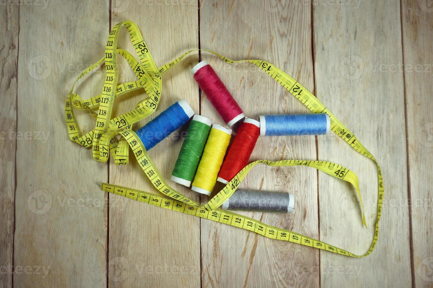 Spools of thread and basic sewing tools including pins, needle, a thimble, a tape measure and yarn scissors on a wooden table photo