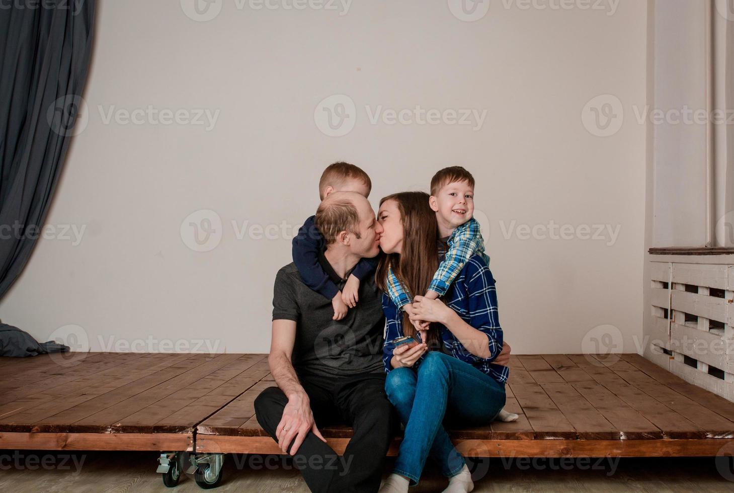 Happy family with two children, in casual clothes, are photographed sitting on the floor. Photoshoot of a family with 2 sons, white background. Warm family relationships. Mom and Dad kiss photo