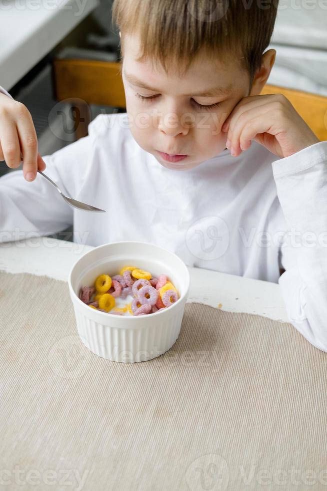 child eats colored cereals at the table, in a white kitchen. Boy's breakfast. Breakfast cereals before school. A quick snack of a schoolboy in the morning. photo