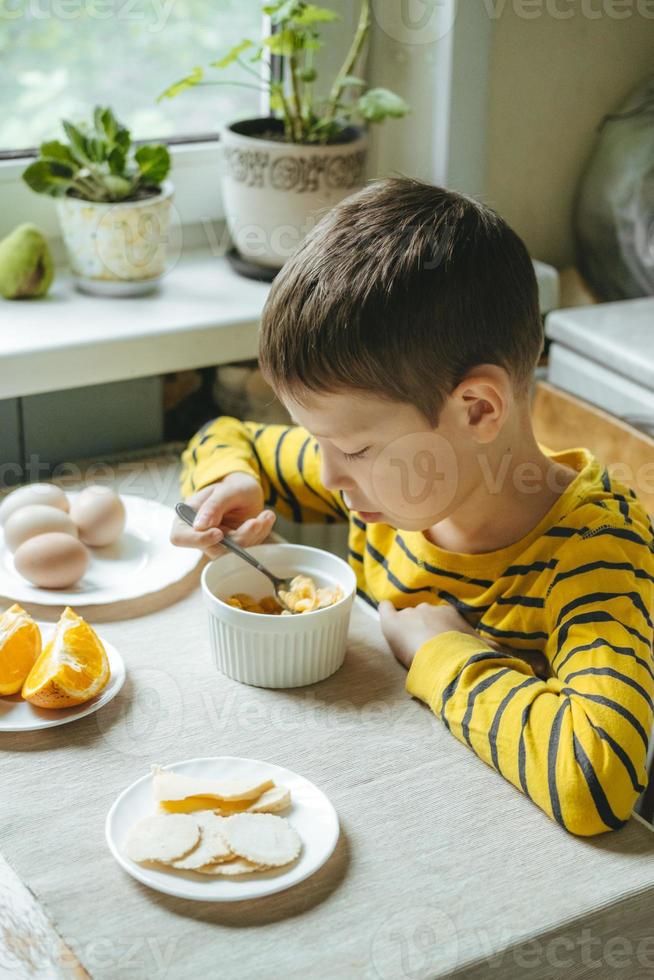 boy eats breakfast in the morning with cereals with milk. Morning breakfast in the kitchen before school. The boy is eating at the table by the window. photo