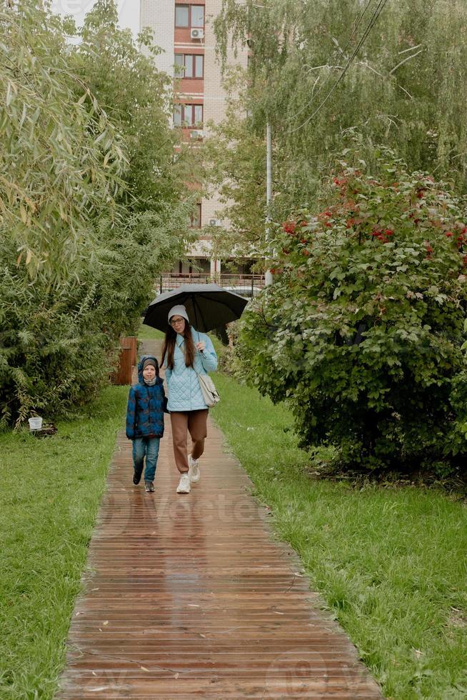 Mom in a blue jacket walks in the park with her son along a wooden path with an umbrella photo