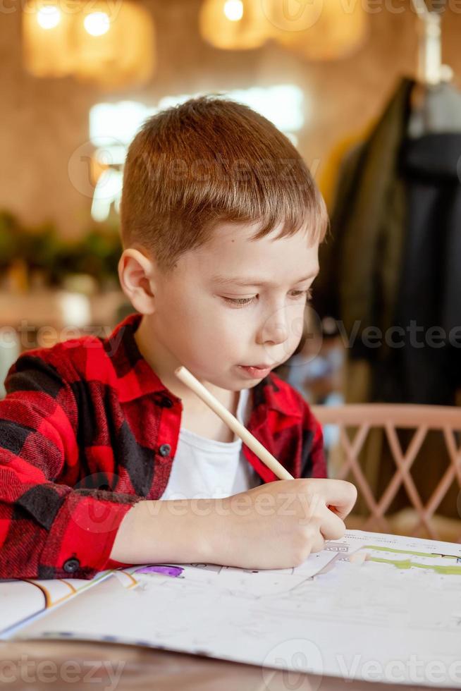 handsome little boy draws at the table with a pencil. Waiting for an order in a cafe. The boy draws on coloring pages in a cafe. photo