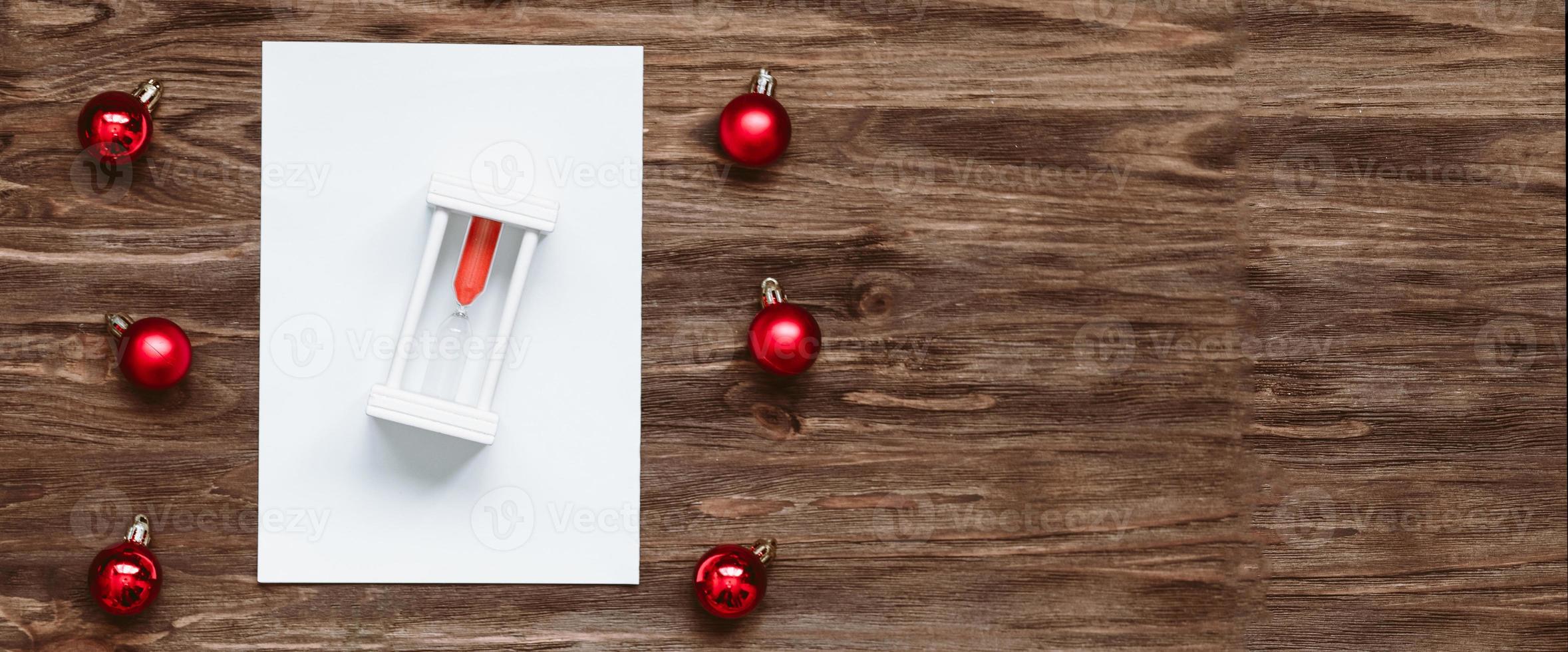 banner hourglass on a blank sheet of paper and and red christmas balls on a wooden background. Top view, flat lay photo