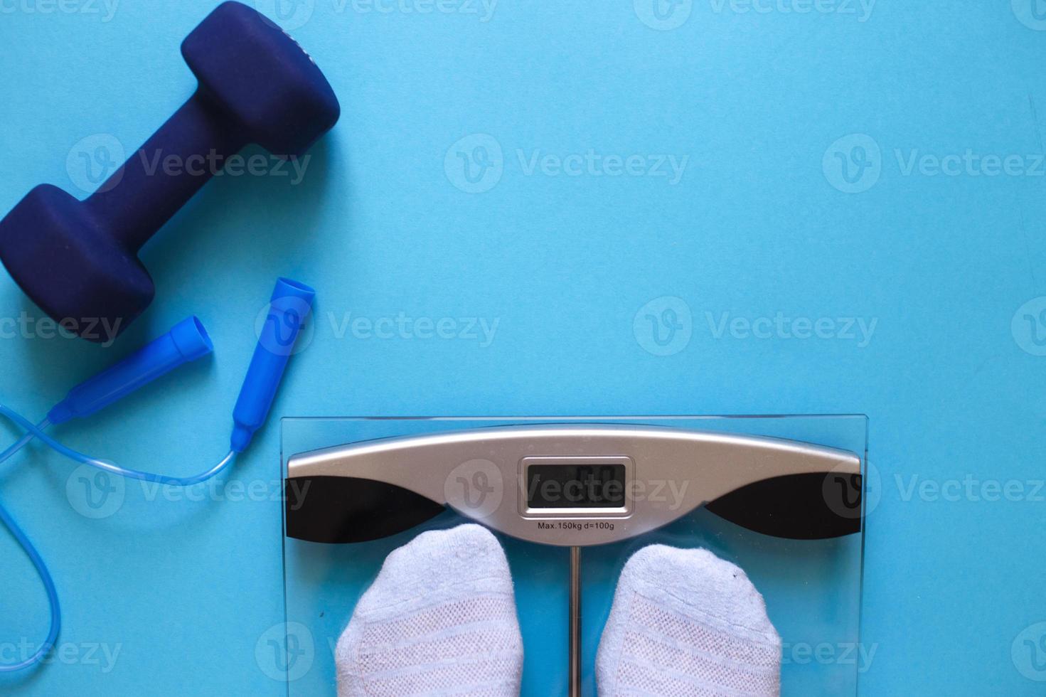 Woman measures her weight on scale on a blue background with skipping rope and dumbbells. Healthy lifestyle accessories photo
