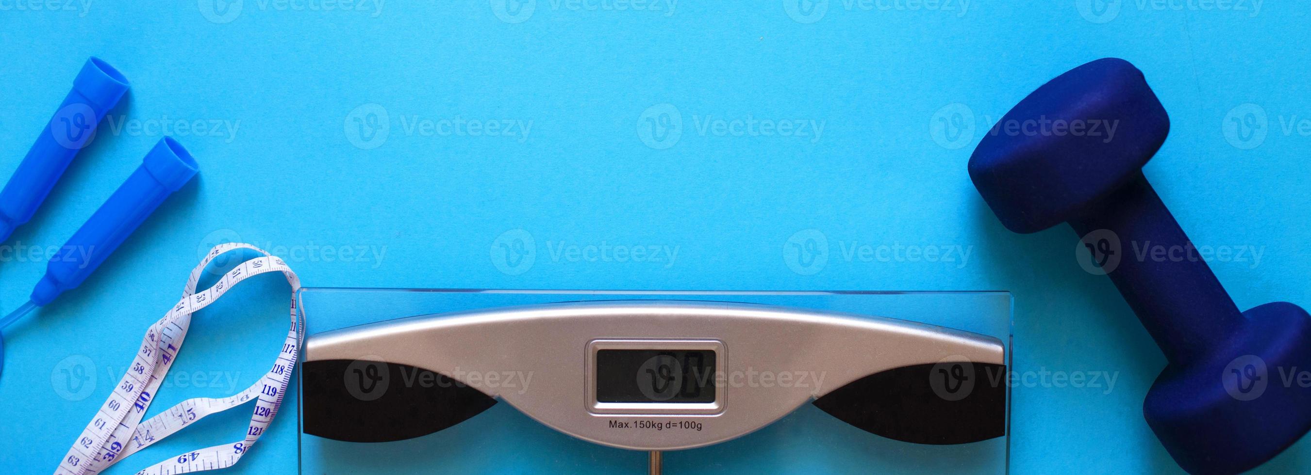 Woman measures her weight on scale on a blue background with skipping rope and dumbbells. Healthy lifestyle banner photo