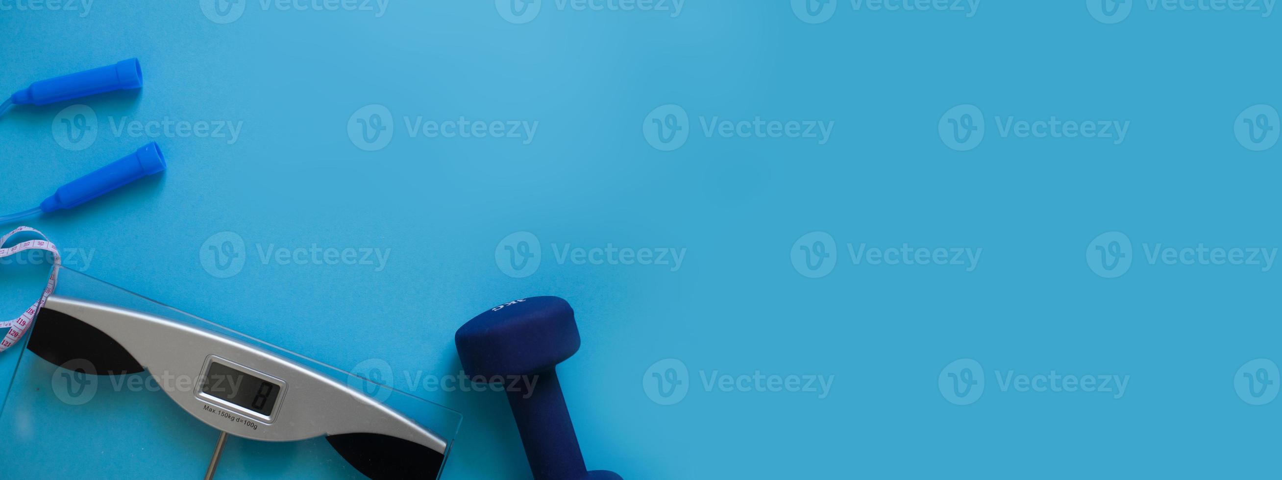 Woman measures her weight on scale on a blue background with skipping rope and dumbbells. Healthy lifestyle banner photo