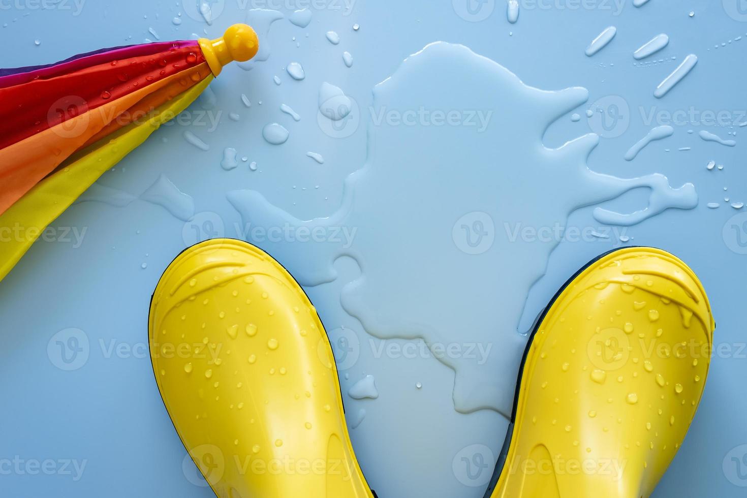 Yellow waterproof boots and a umbrella on a blue background in a puddle of water. Concept of preparation for the rainy season. Copy space. photo