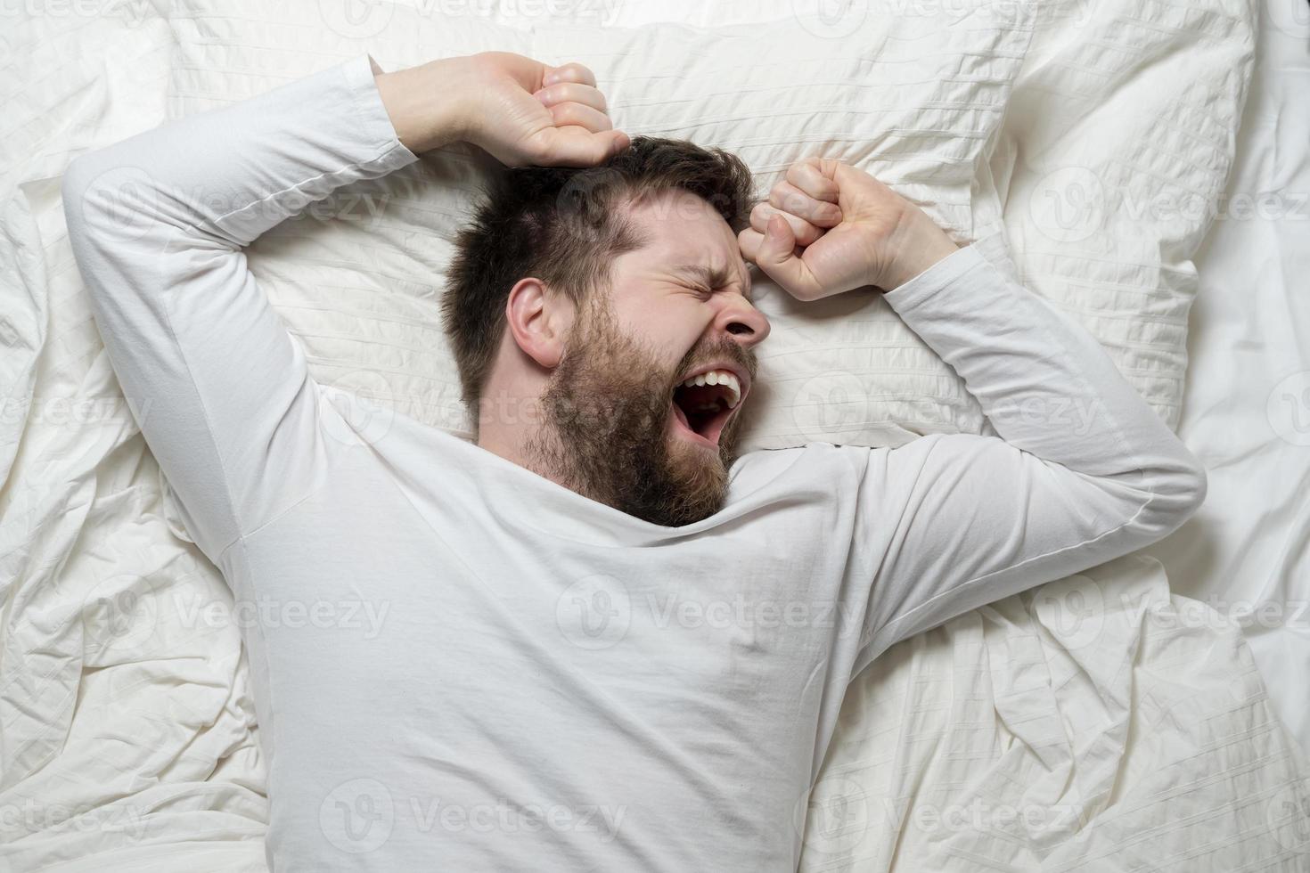 Bearded man in white pajamas is tired and wants to sleep, he yawns and stretches while lying in bed. Top view. Close-up. photo