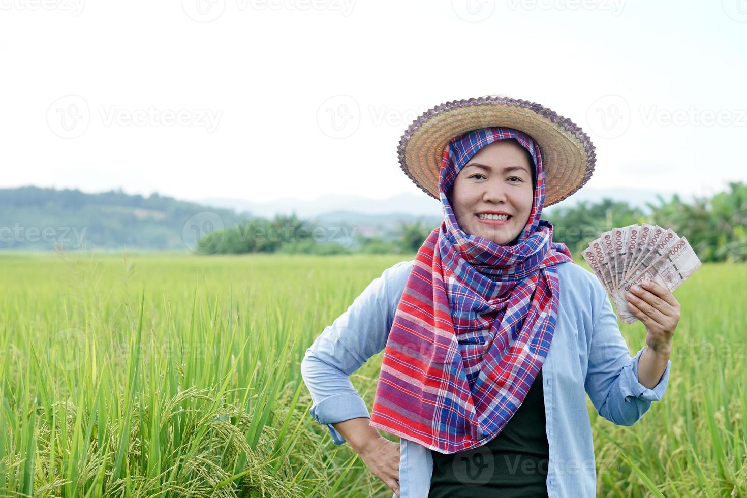 Asian farmer woman is at paddy field, hold Thai banknote money. Concept , Farmer happy to get profit, income, agriculture supporting money. Proud on crops. photo