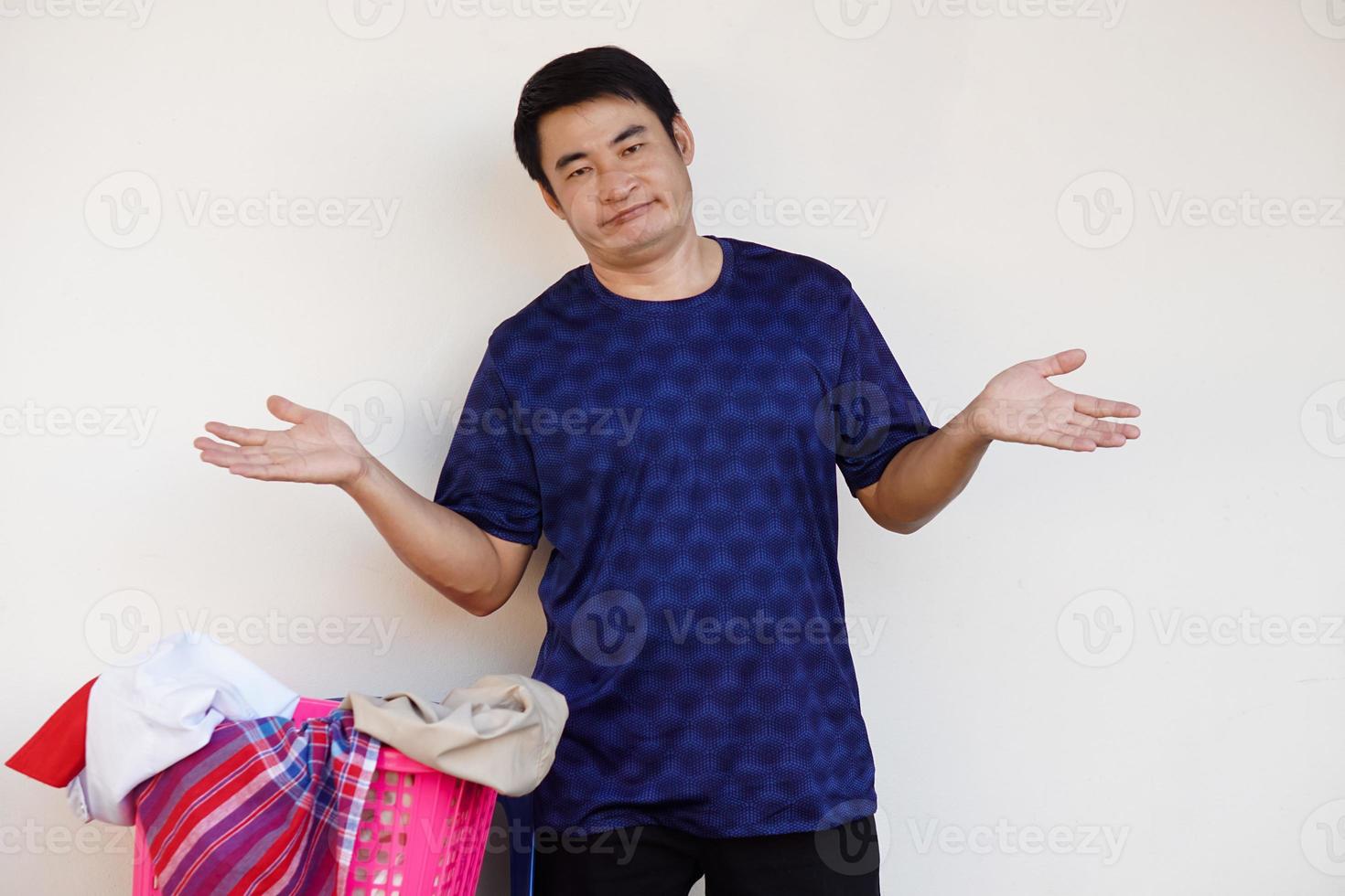 el hombre asiático se para al lado de la canasta de ropa, pone las manos en las caderas, sintiendo que no quiere lavar la ropa. concepto, tarea aburrida, tareas domésticas. foto