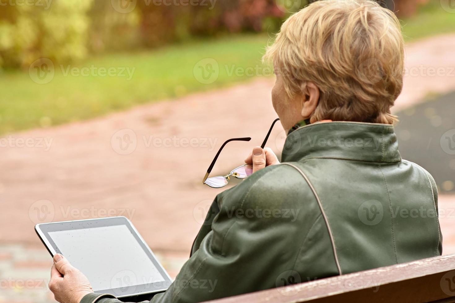 an elderly woman with a tablet computer is sitting on a bench in an autumn park. photo
