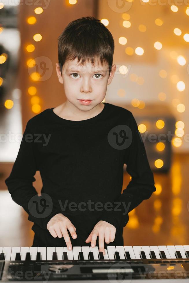 a boy in a black T-shirt plays the piano, against the background of a Christmas garland photo