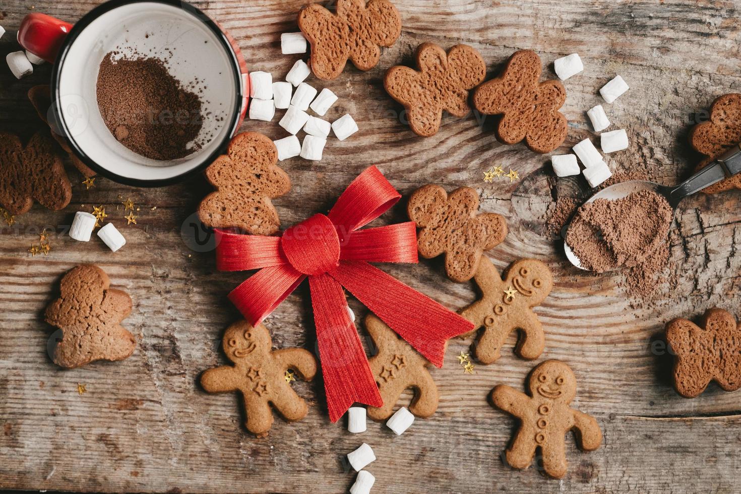 hombre de pan de jengibre, cacao y galletas con un gran lazo rojo sobre un fondo de madera, vista superior. varios dulces navideños con una taza de cacao en la mesa foto