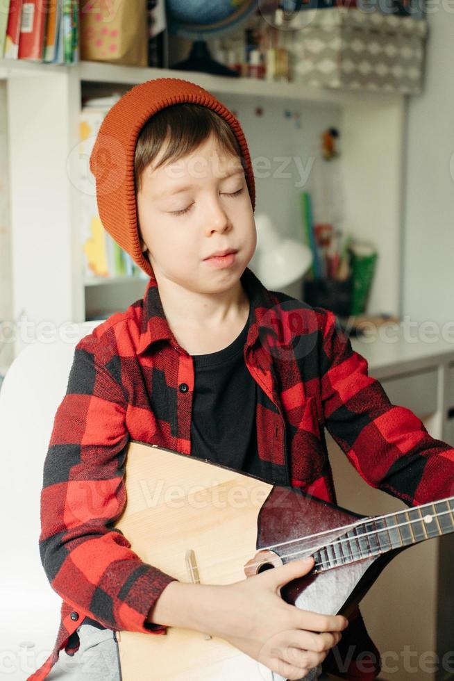 niño con un sombrero rojo y una camisa a cuadros toca la balalaika. chico guapo sosteniendo su guitarra. clases de música en casa. pasatiempo para el alma. música de enseñanza en casa foto