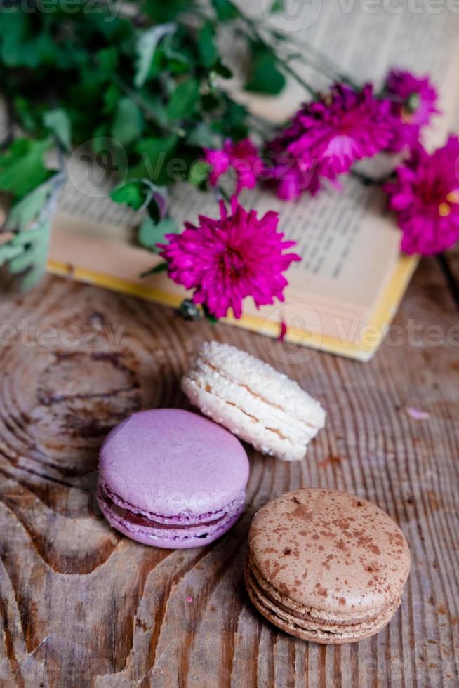 macarons fondo flores rojas y libros, sobre una mesa de madera. marco vertical. estética con macarrones y flores. hermosos pasteles en una mesa de madera. desayuno francés por la mañana. foto