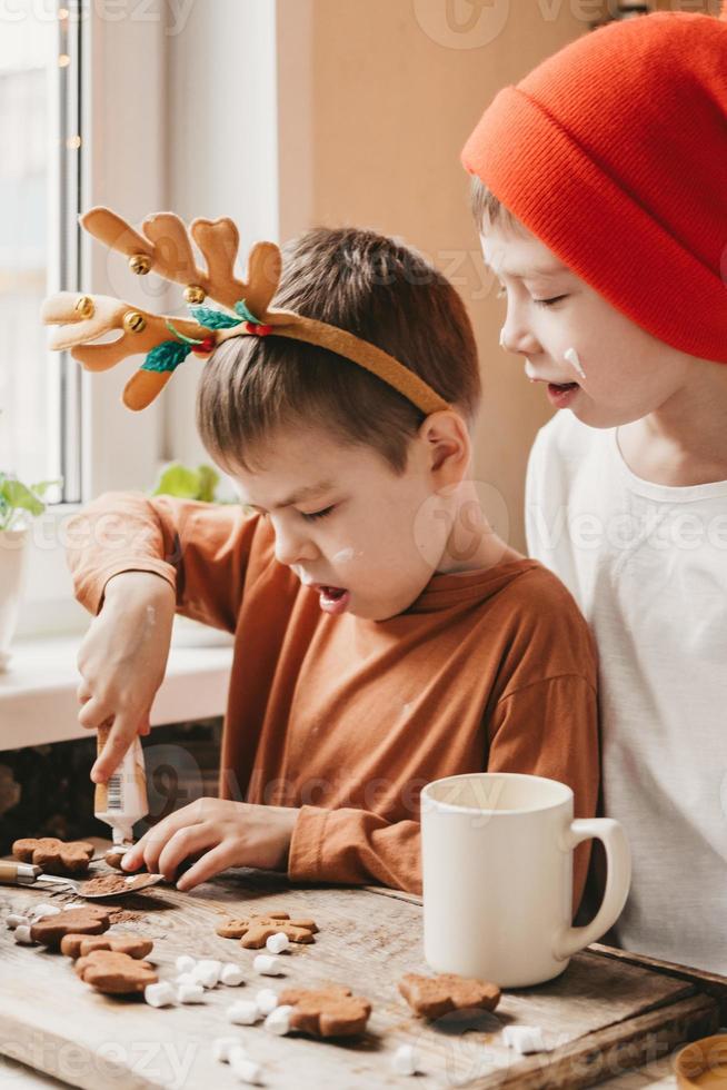 Boys decorate Christmas cookies with white icing. Children decorate a gingerbread man on a wooden background. Christmas sweets with cocoa and marshmallows for children. photo