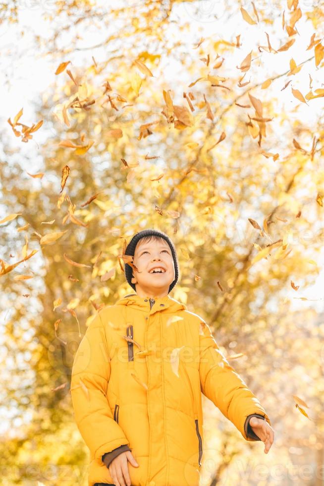 niño con una chaqueta amarilla, esparce hojas en un parque de otoño. el niño se regocija en las hojas de otoño. infancia feliz. chaqueta amarilla brillante y hojas foto