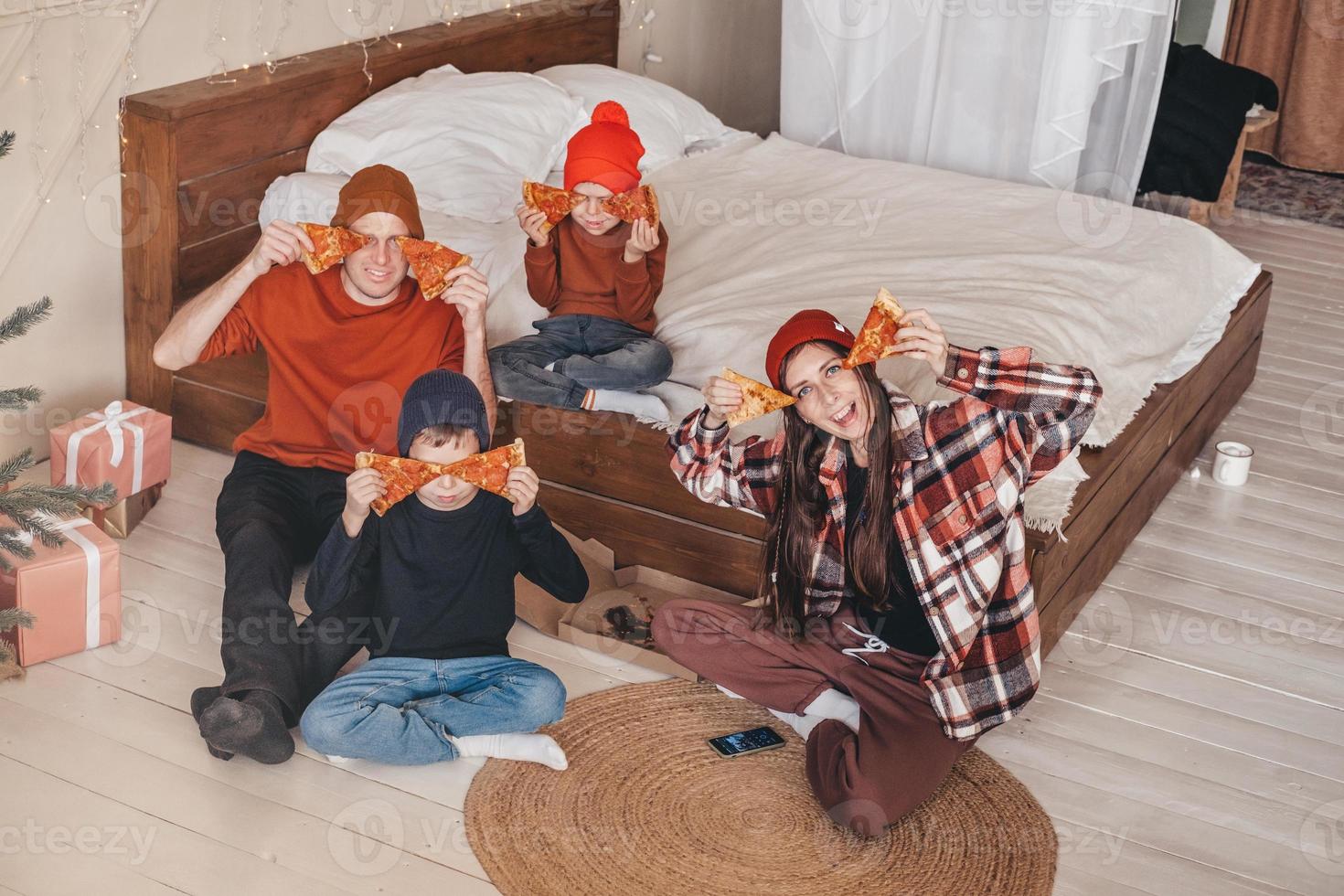 funny family photo with two children eating pizza in bed. happy family moment on the weekend. A family with two children is eating an ordered pizza from a box in the bedroom. Boho style bedroom