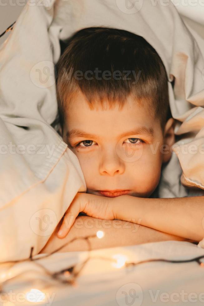 portrait of a serious boy lying under a white blanket and with a garland. child's christmas morning. Boy wake up in the morning, vertical frame photo