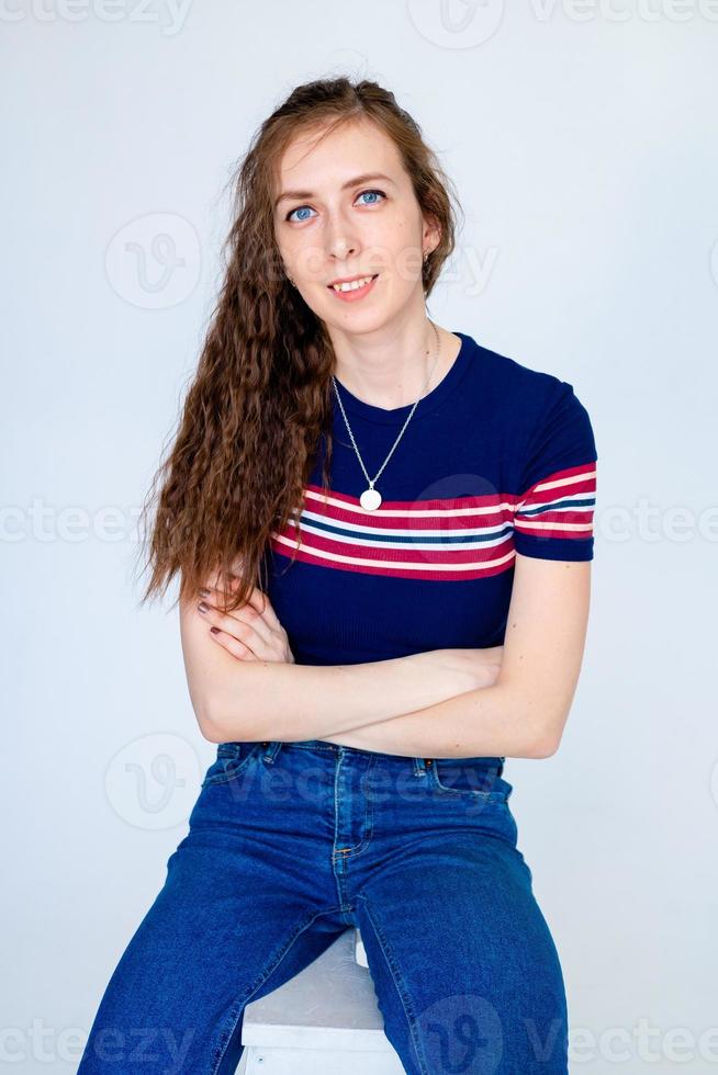 portrait of a young smiling girl sitting on a chair, on a white background, with long hair, in a tight T-shirt and jeans photo