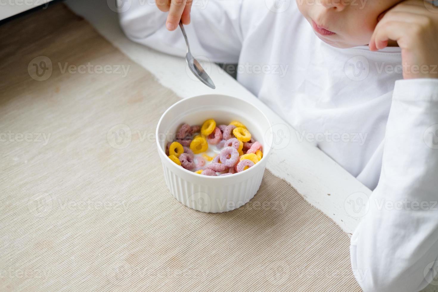 child eats colored cereals at the table, in a white kitchen. Boy's breakfast. Breakfast cereals before school. A quick snack of a schoolboy in the morning. photo
