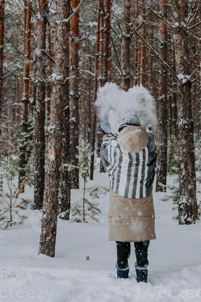 Tossed snow in the form of a heart in the air. A girl in a striped scarf stands in a winter forest and throws snow in the air. A girl stands with her back in the woods in a wide striped scarf. photo