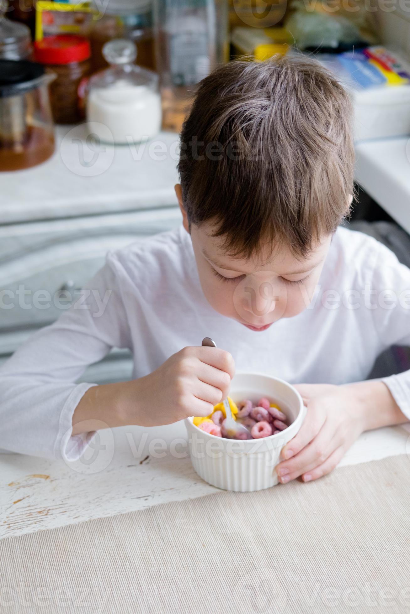 el niño come cereales de colores en la mesa, en una cocina blanca