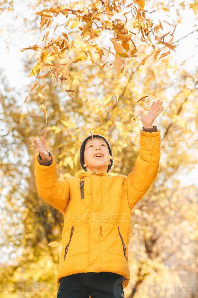 niño con una chaqueta amarilla, esparce hojas en un parque de otoño. el niño se regocija en las hojas de otoño. infancia feliz. chaqueta amarilla brillante y hojas foto