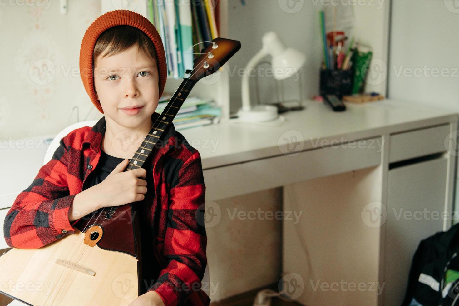 niño con un sombrero rojo y una camisa a cuadros toca la balalaika. chico guapo sosteniendo su guitarra. clases de música en casa. pasatiempo para el alma. música de enseñanza en casa foto