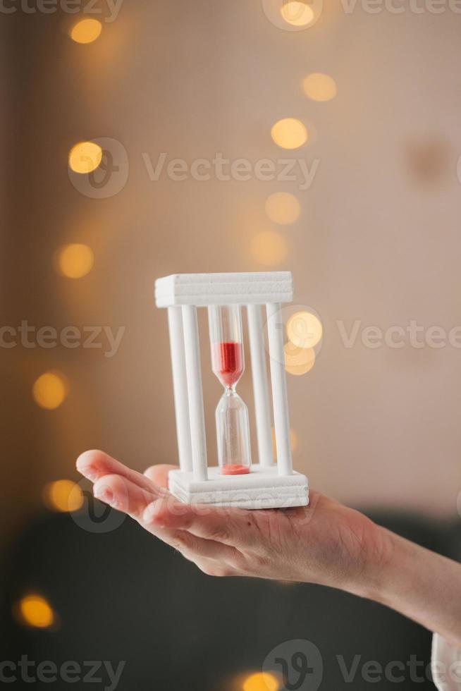 hands hold a white hourglass on the background of a Christmas garland. Hourglass close-up. Time is running. Red sand in hours. New Years is soon. Time for a change photo