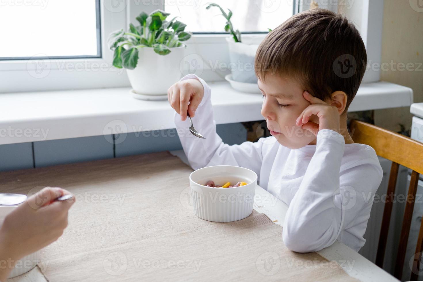 child eats colored cereals at the table, in a white kitchen. Boy's breakfast. Breakfast cereals before school. A quick snack of a schoolboy in the morning. photo