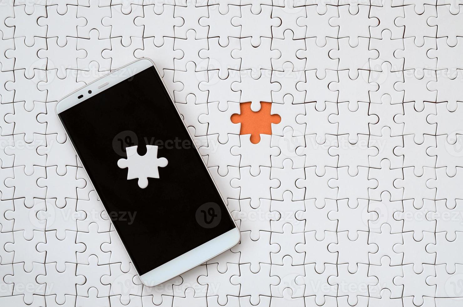 A modern big smartphone with several puzzle elements on the touch screen lies on a white jigsaw puzzle in an assembled state with missing elements photo