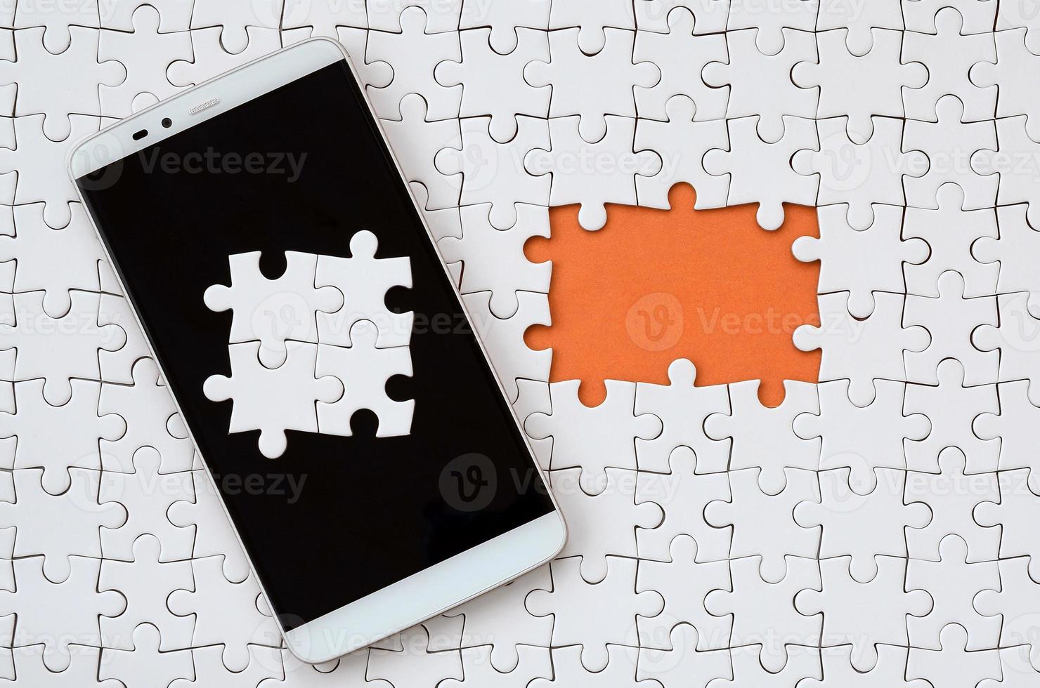 A modern big smartphone with several puzzle elements on the touch screen lies on a white jigsaw puzzle in an assembled state with missing elements photo
