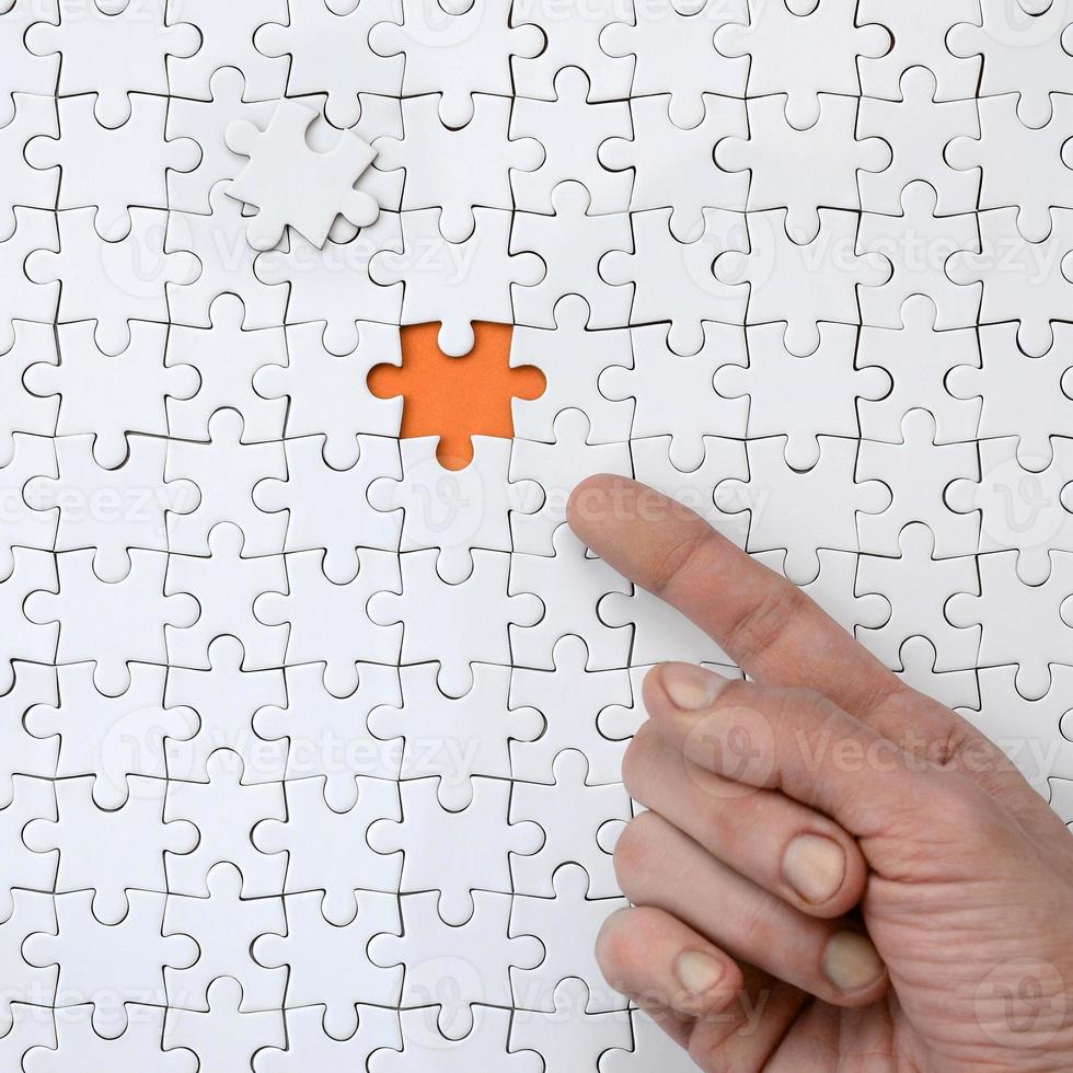 The texture of a white jigsaw puzzle in the assembled state with one missing element, forming an orange space, pointed to by the finger of the male hand photo