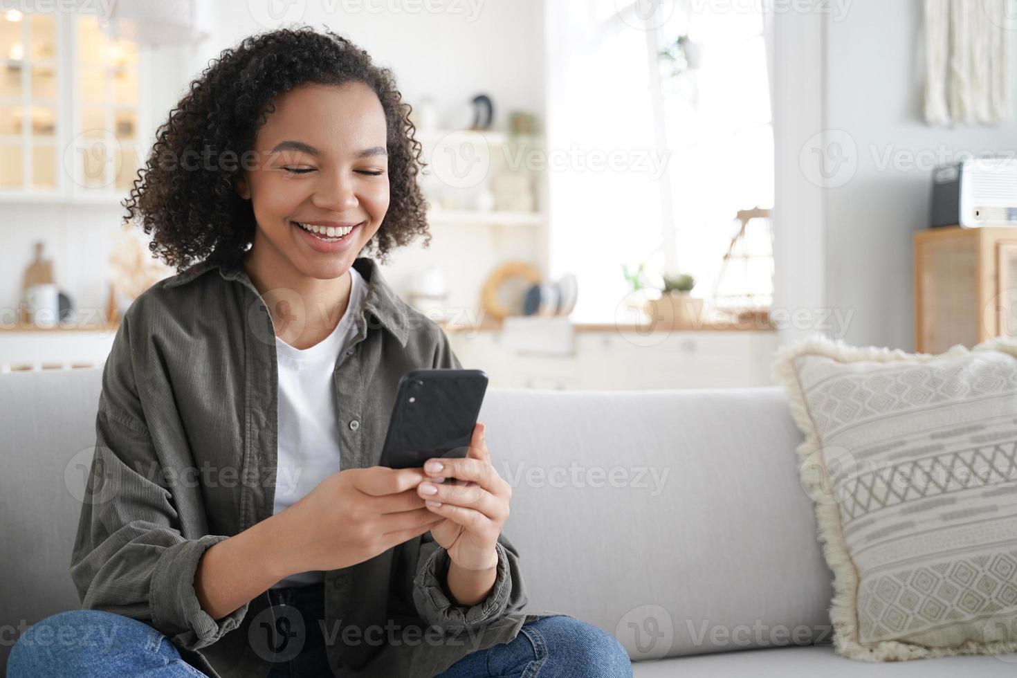 Happy mixed race girl uses mobile phone apps watching funny video in social networks sitting on sofa photo