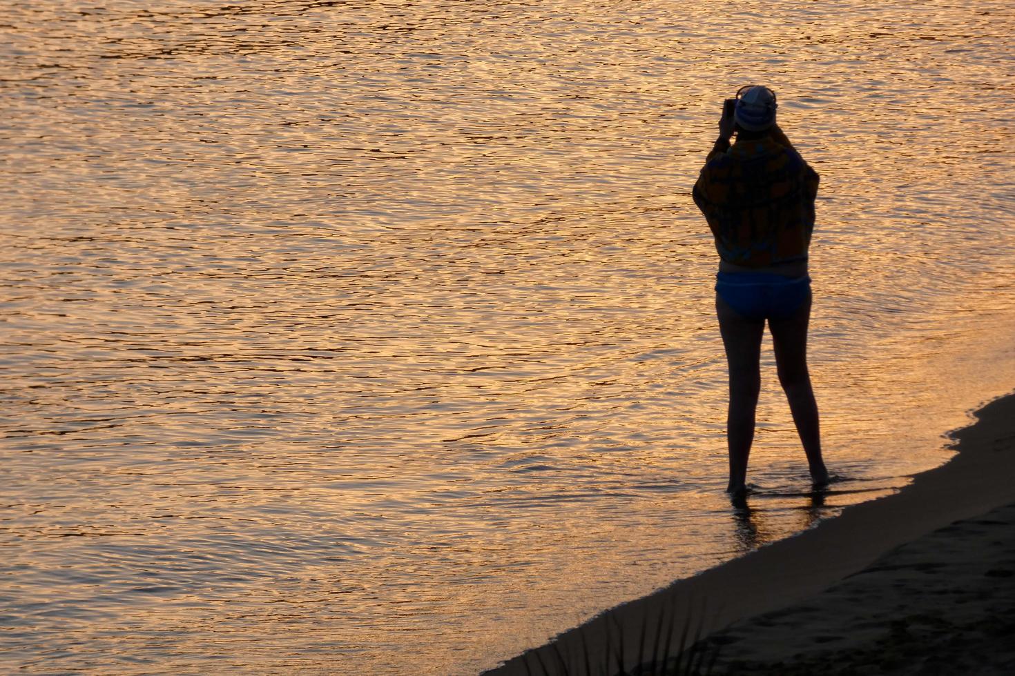 Backlight of a silhouette of an anonymous woman taking photos in the sea