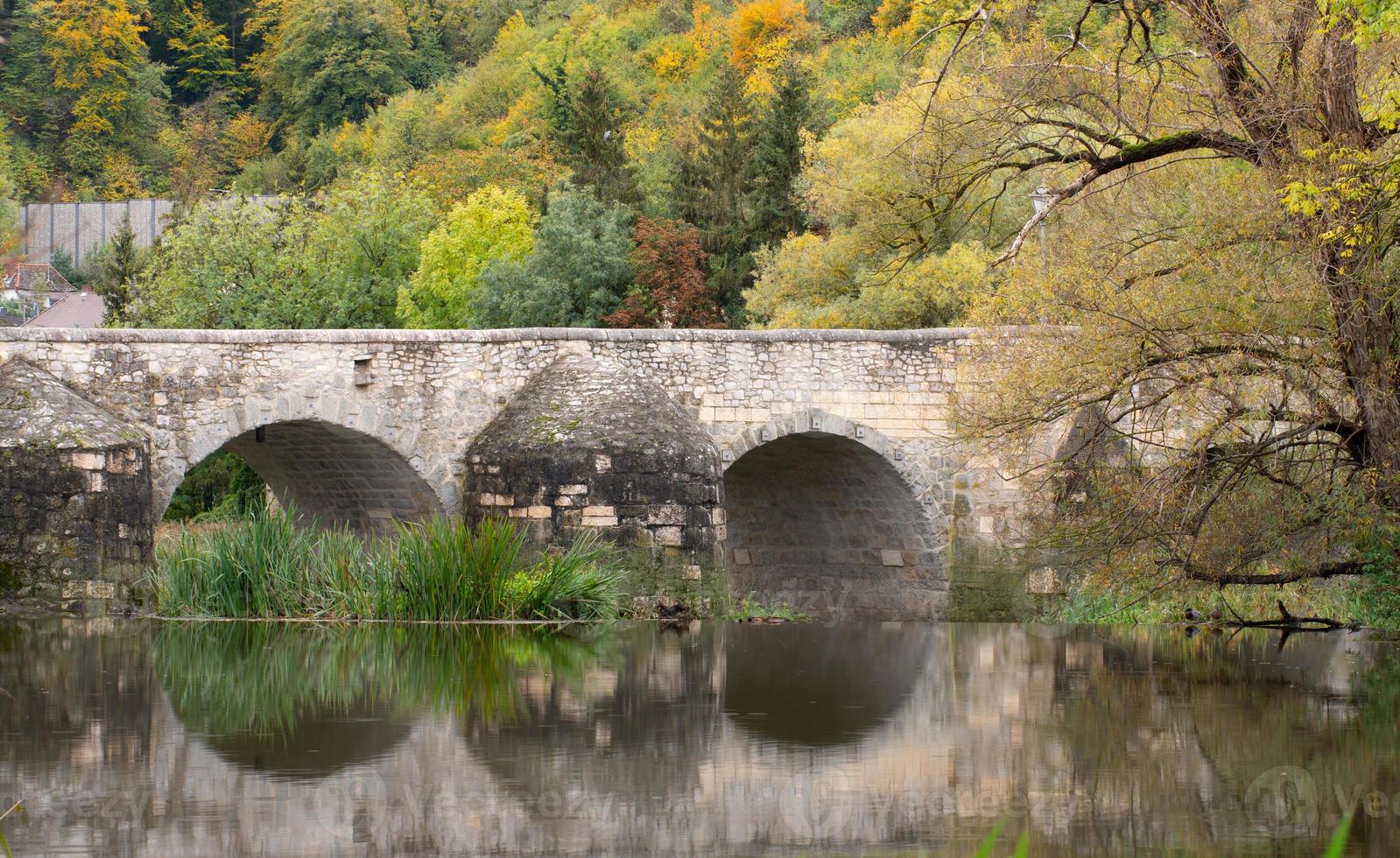 A historic stone bridge, with several round arches, is reflected in the water. In the background deciduous trees in autumn with golden leaves. photo