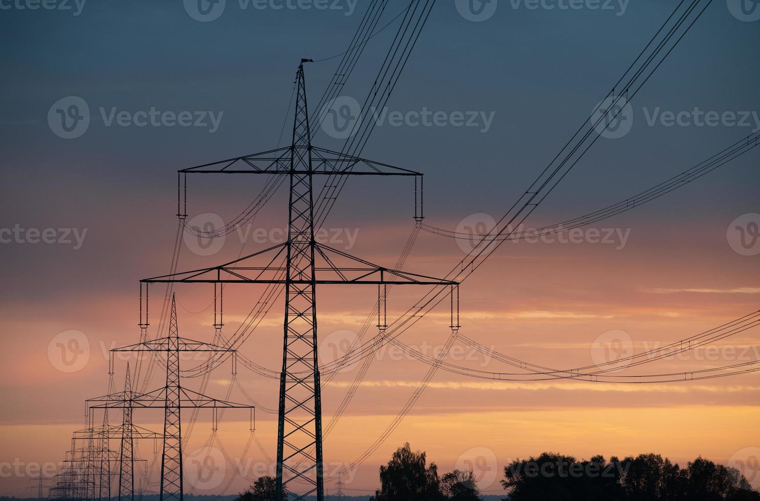 Close-up of large overhead power lines carrying electricity over long distances. The sun rises and shines in the background. photo