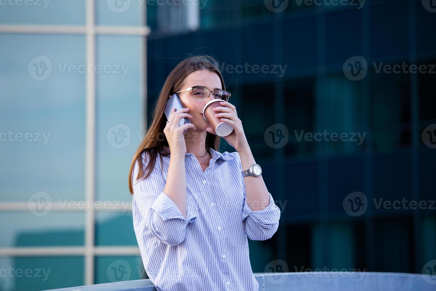 Executive business woman talking on mobile smartphone and drinking coffee from disposable paper cup in the street with office buildings in the background photo