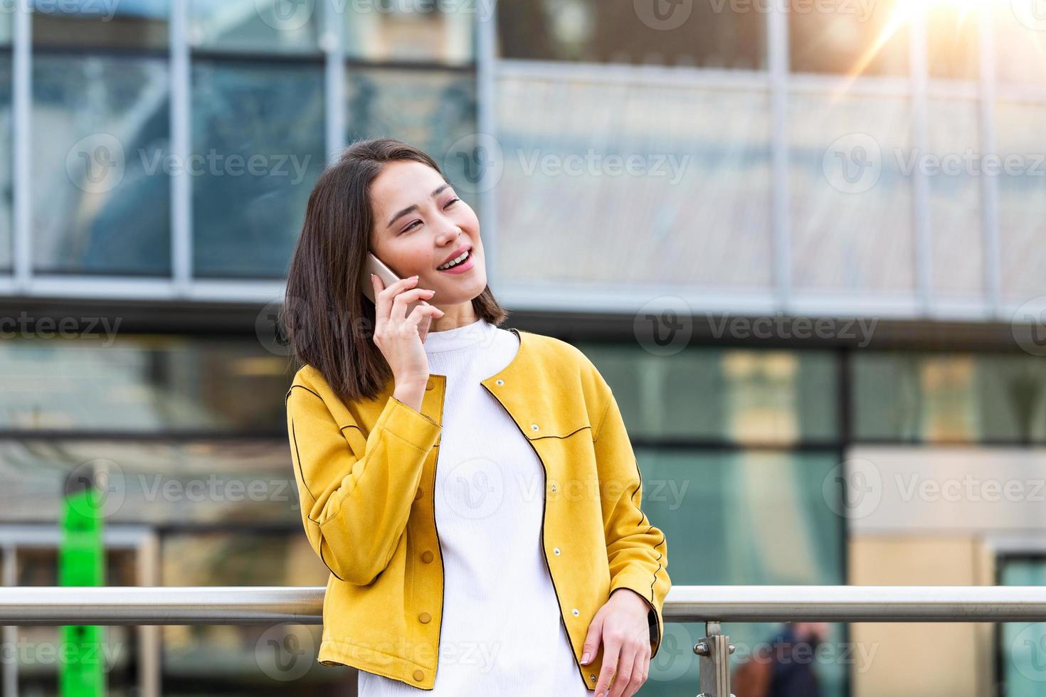 Happy smiling business woman talking on mobile smartphone in the street with office buildings in the background. Executive business Asian woman talking on mobile smartphone photo