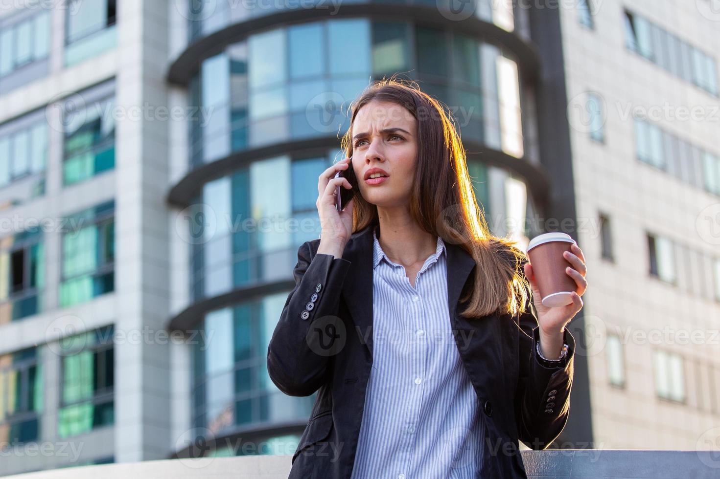 Business woman with coffee arguing on the phone over bad business decitions. Young woman with smartphone standing against street blurred building background. photo