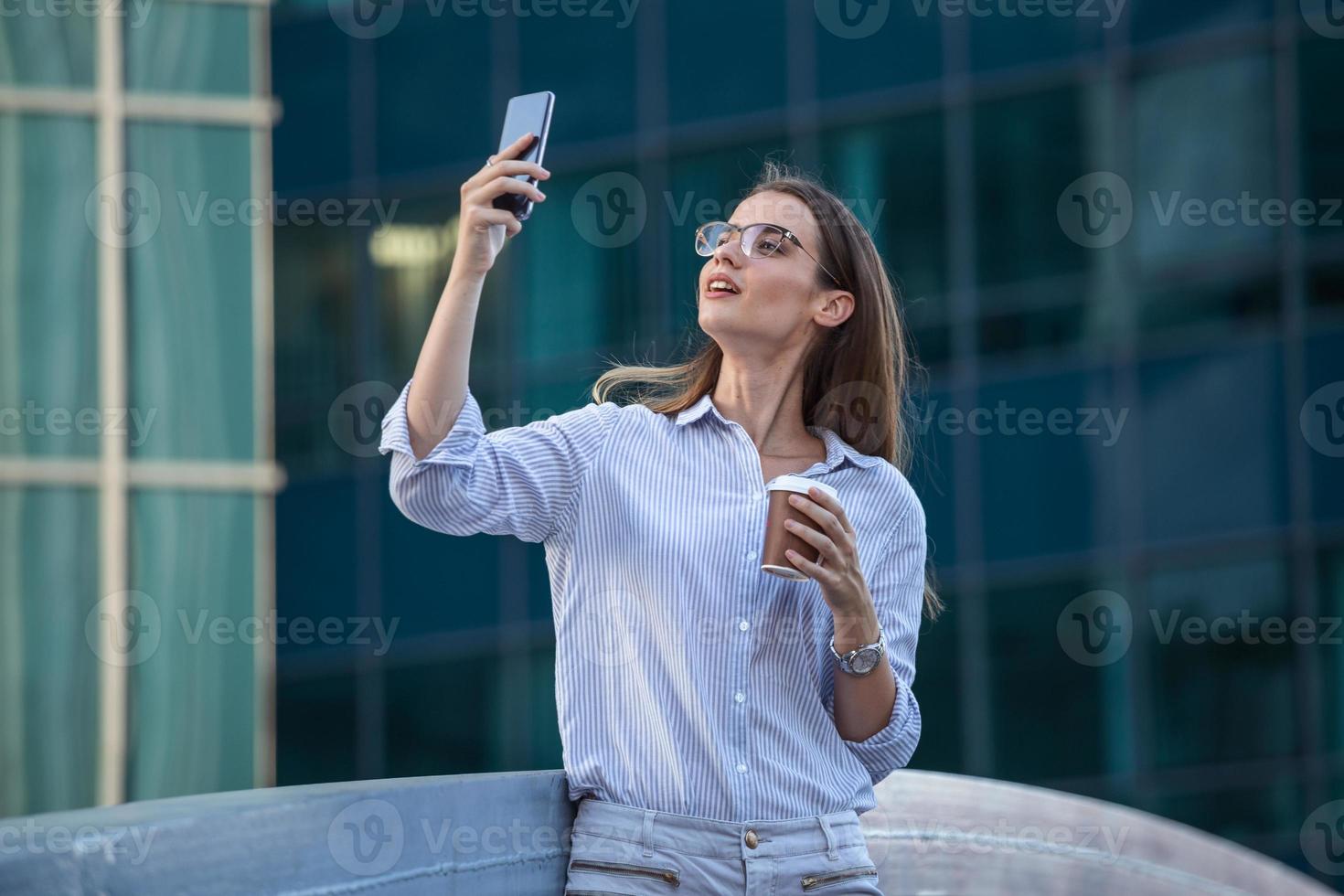 Executive business woman looking at mobile smartphone and drinking coffee from disposable paper cup in the street with office buildings in the background photo