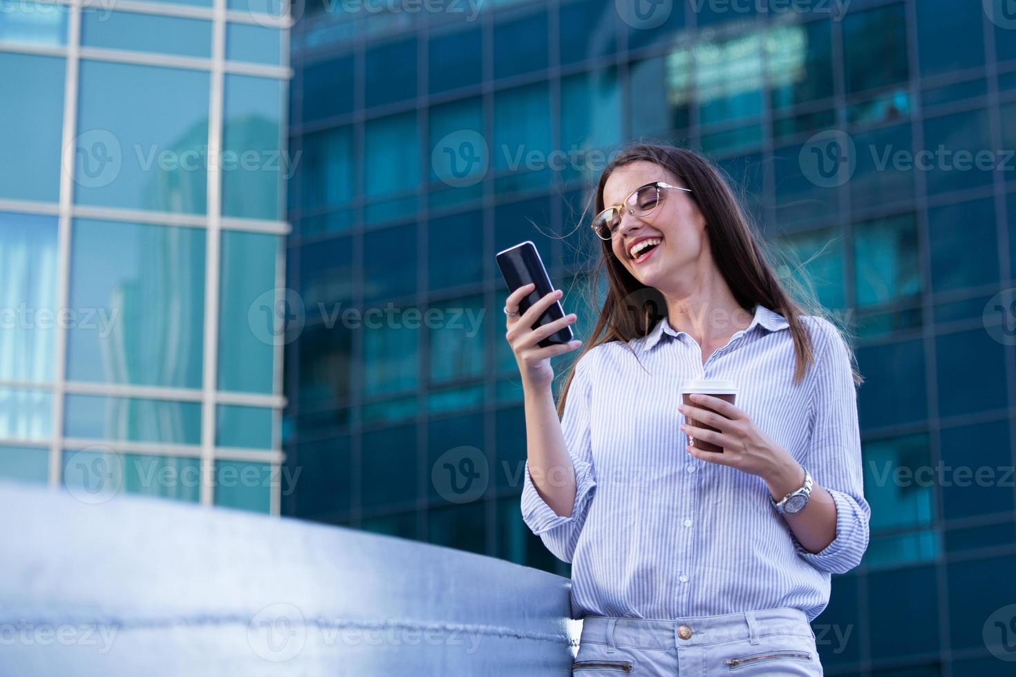 Executive business woman looking at mobile smartphone and drinking coffee from disposable paper cup in the street with office buildings in the background photo