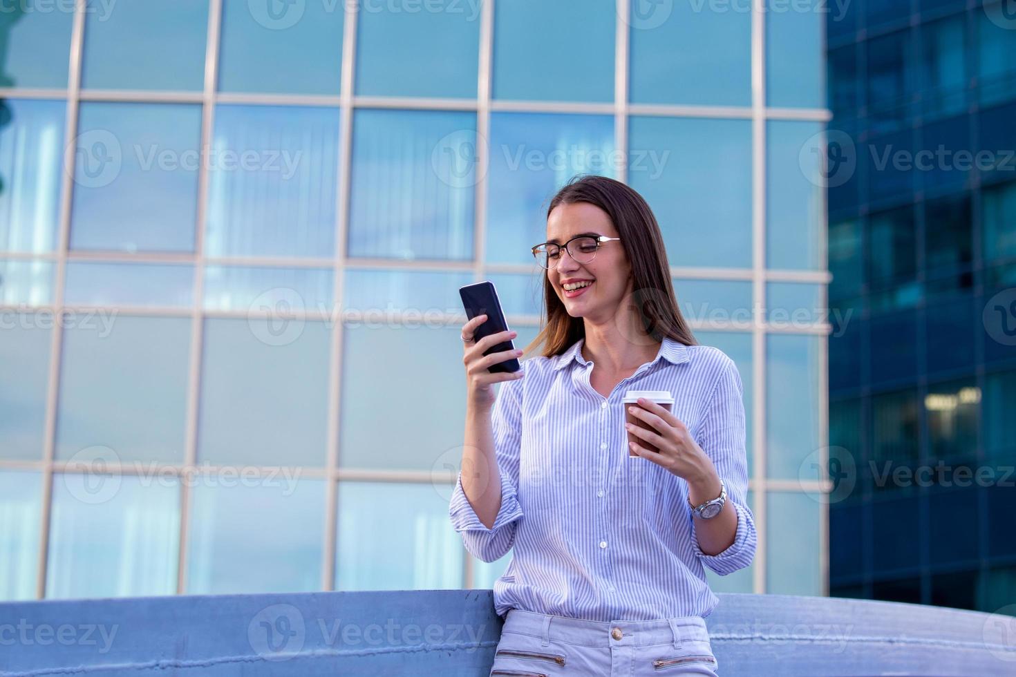 Executive business woman looking at mobile smartphone and drinking coffee from disposable paper cup in the street with office buildings in the background photo