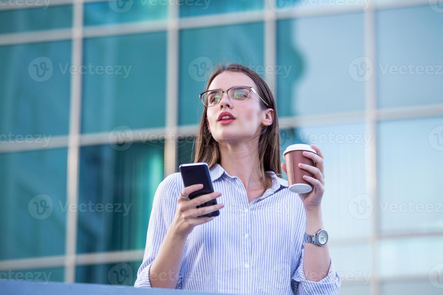 Executive business woman looking at mobile smartphone and drinking coffee from disposable paper cup in the street with office buildings in the background photo