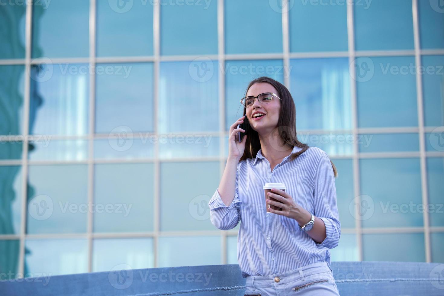 Executive business woman talking on mobile smartphone and drinking coffee from disposable paper cup in the street with office buildings in the background photo