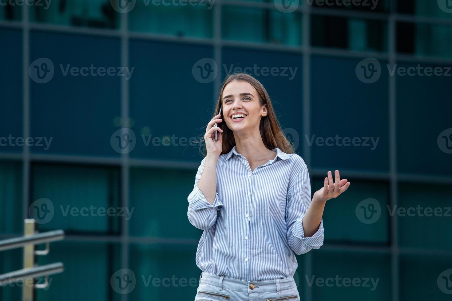Happy smiling business woman talking on mobile smartphone and walking in the street with office buildings in the background photo