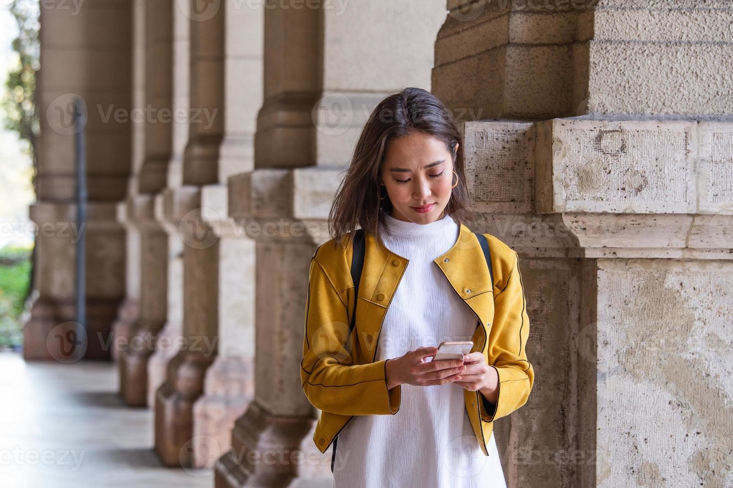 Beautiful asian woman using an application to send an sms message in her smartphone device. Happy young Asian woman with smartphone standing in the street photo